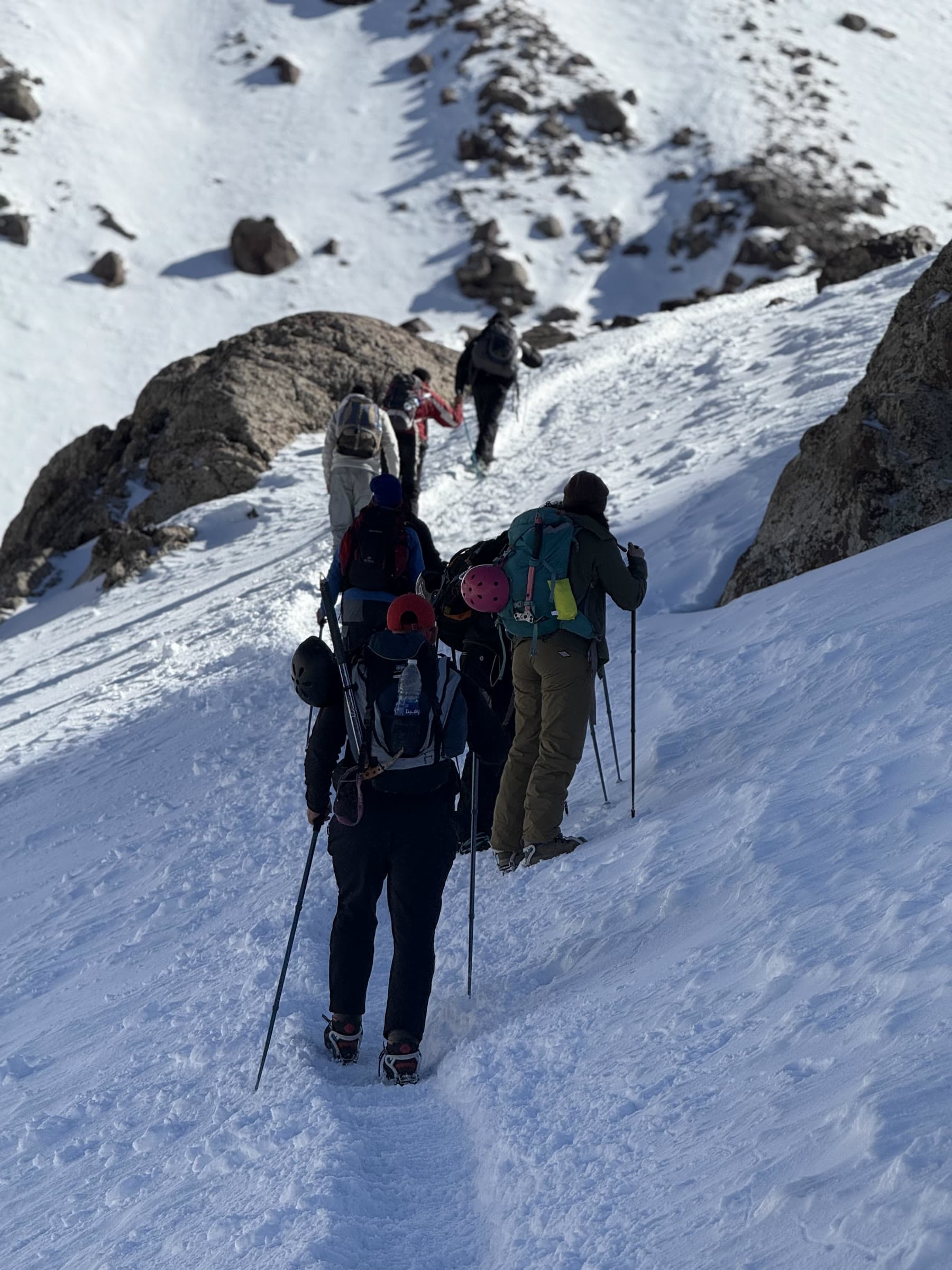 A group climbing a snow slope on crampons with poles, spaced out at their own pace