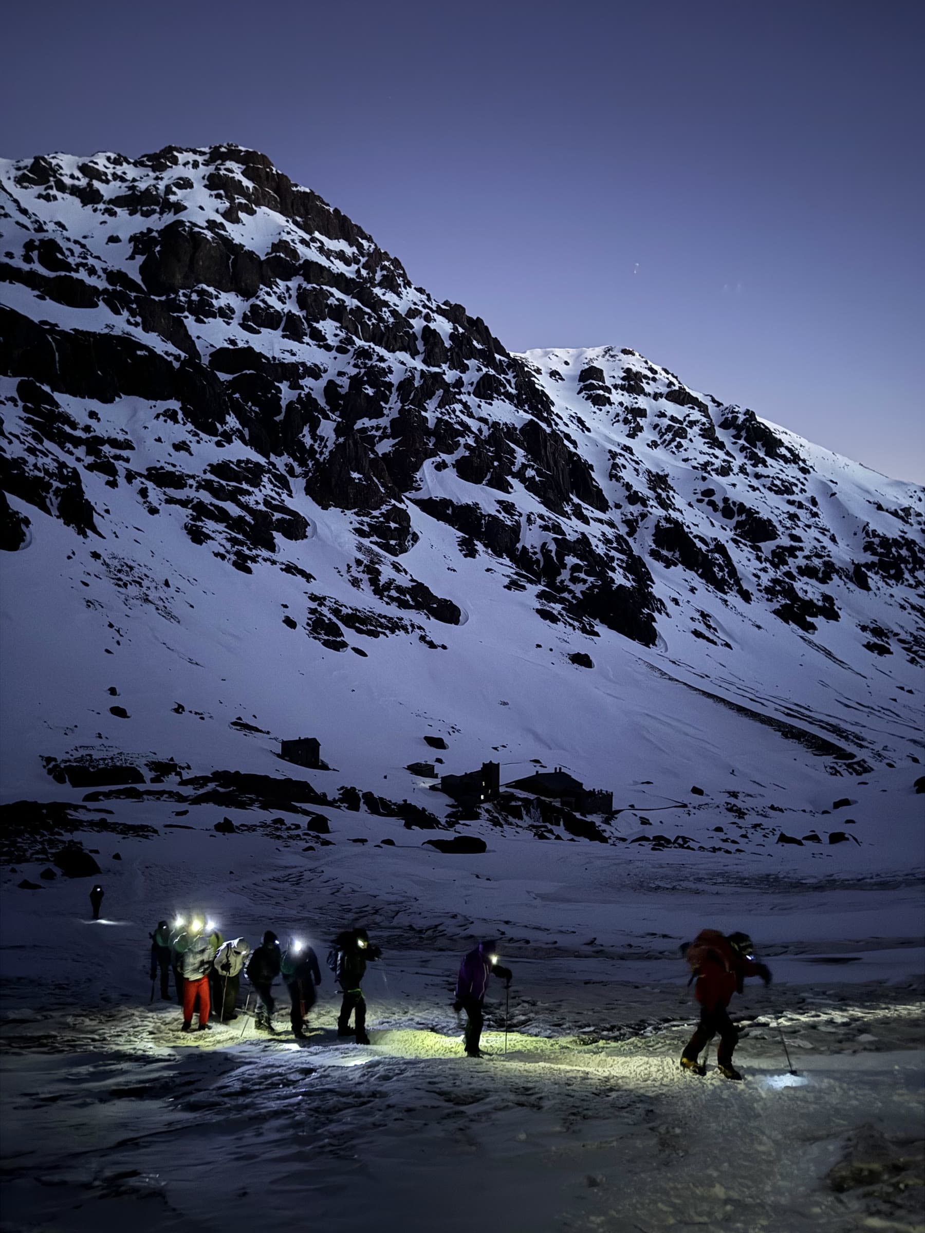 A group setting off across the snow by headtorch in the blue light before dawn, the refuge behind them
