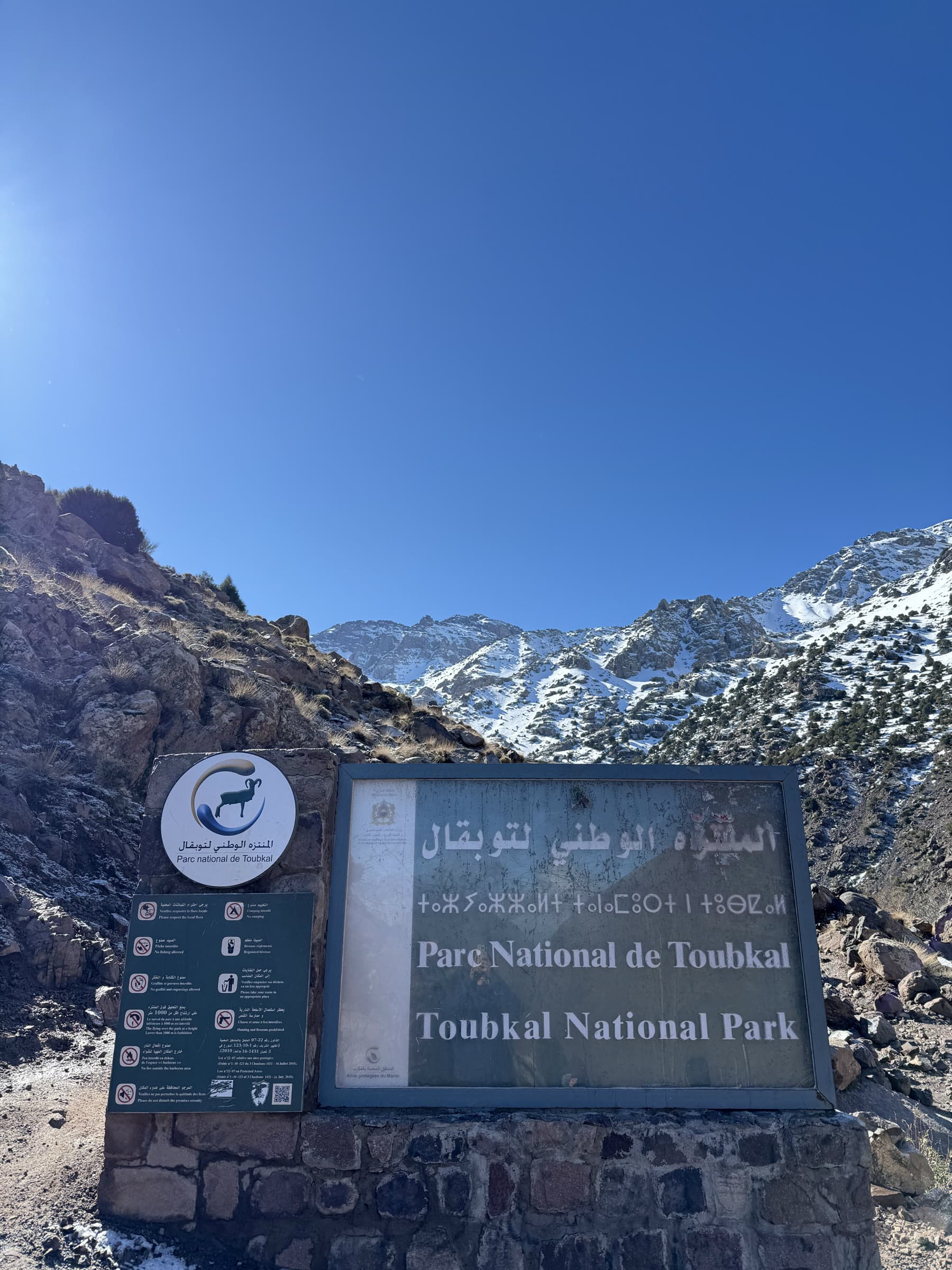 The Toubkal National Park sign in Arabic, Tifinagh, French and English, snow ridges behind it