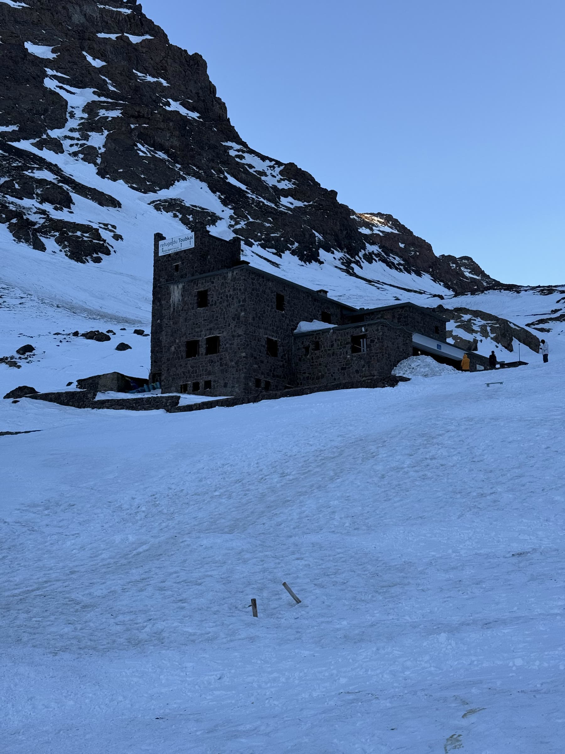 The stone refuge at 3,207 m sitting in old snow beneath a dark rock face
