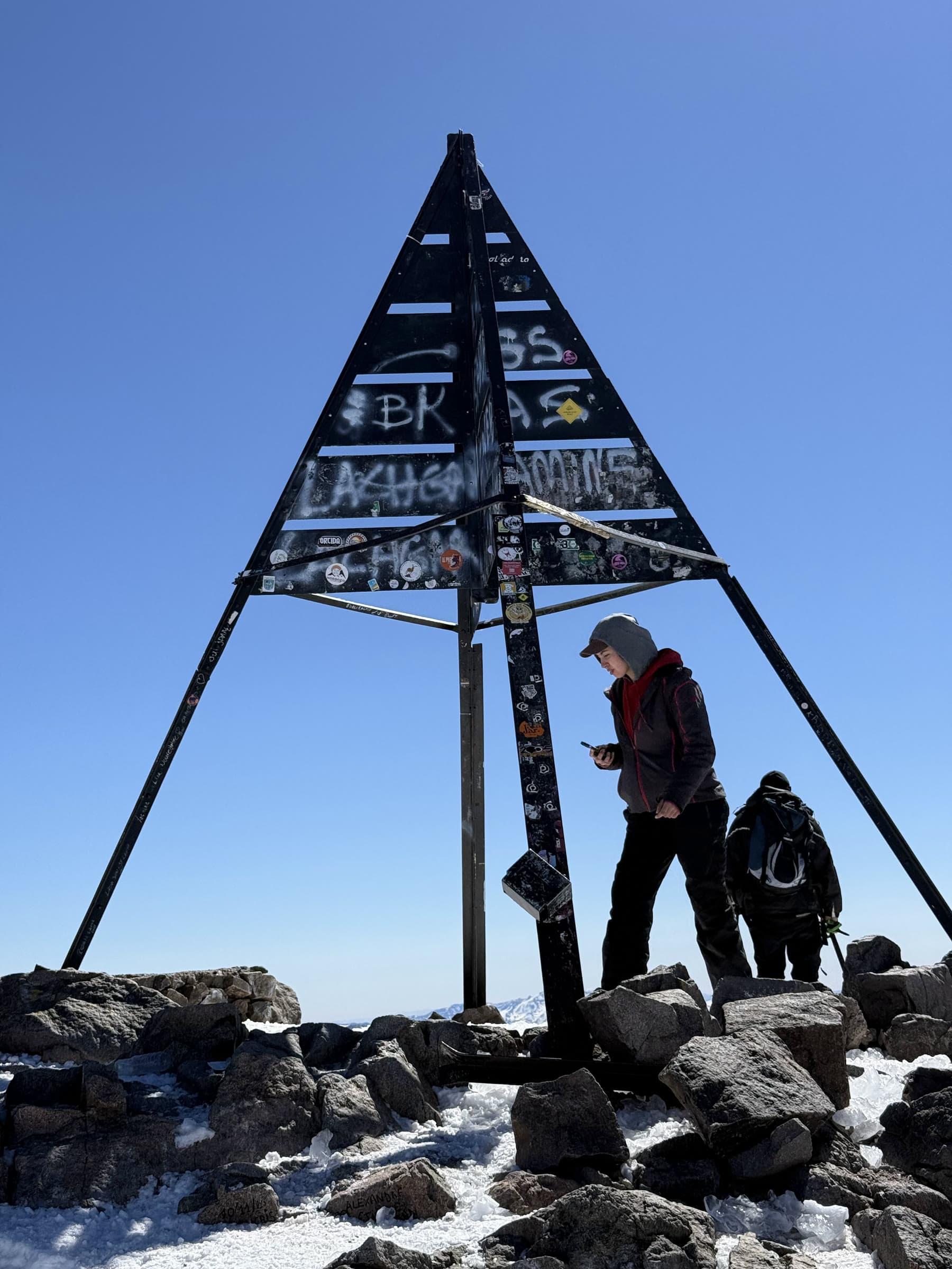 The graffitied steel pyramid at the summit of Toubkal with two walkers beside it