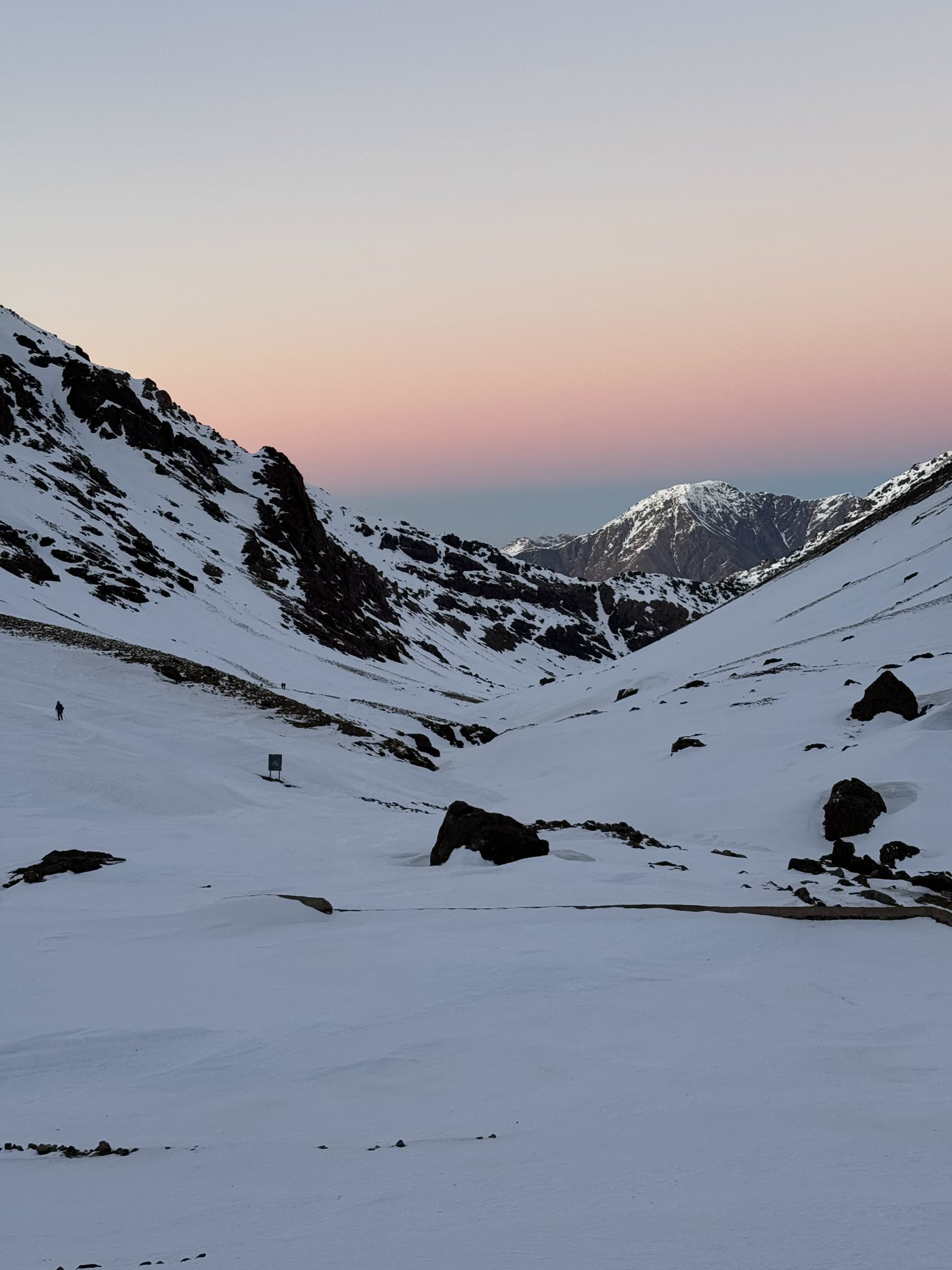 First light turning the sky pink over a snow cwm, a single walker small against the slope