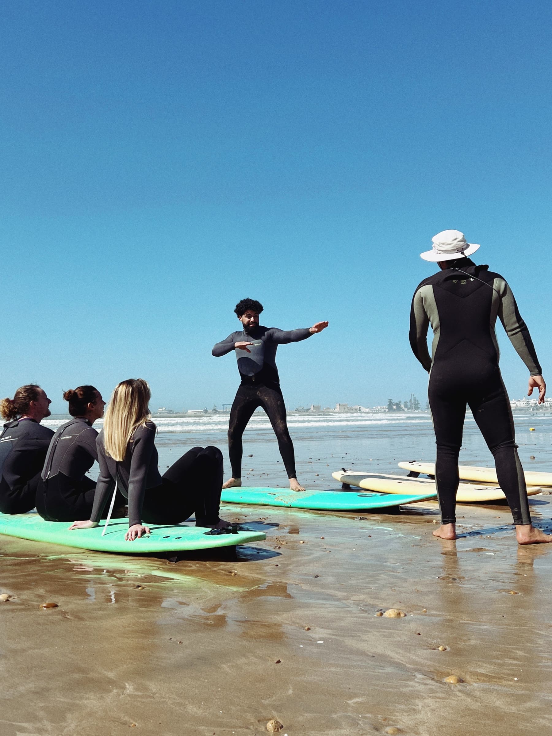 A surf lesson on a wide Atlantic beach: an instructor demonstrating the pop-up, learners waiting on their boards