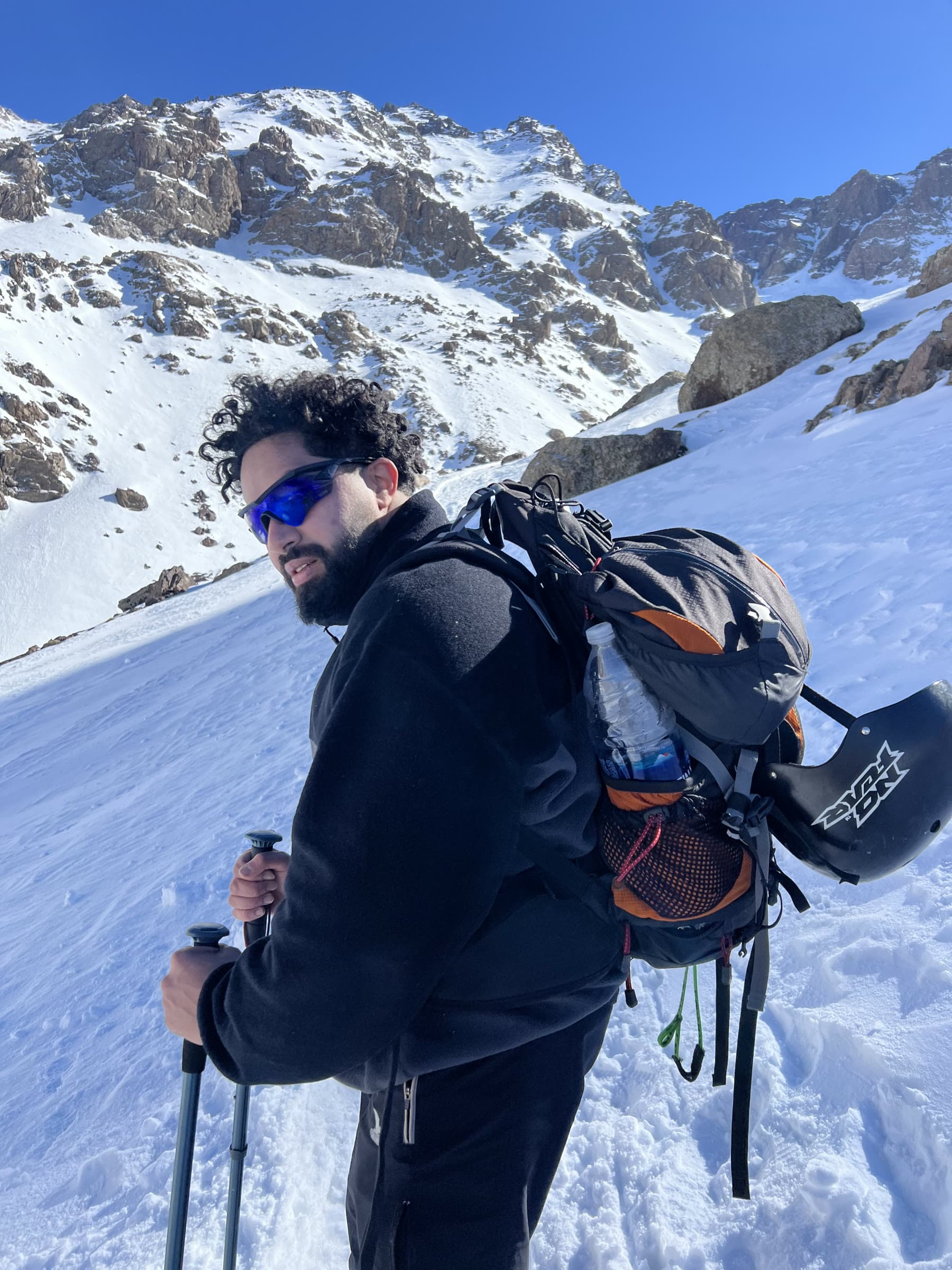 Zakaria Basraoui on the trail, pack on, a snowed rock face filling the frame behind him