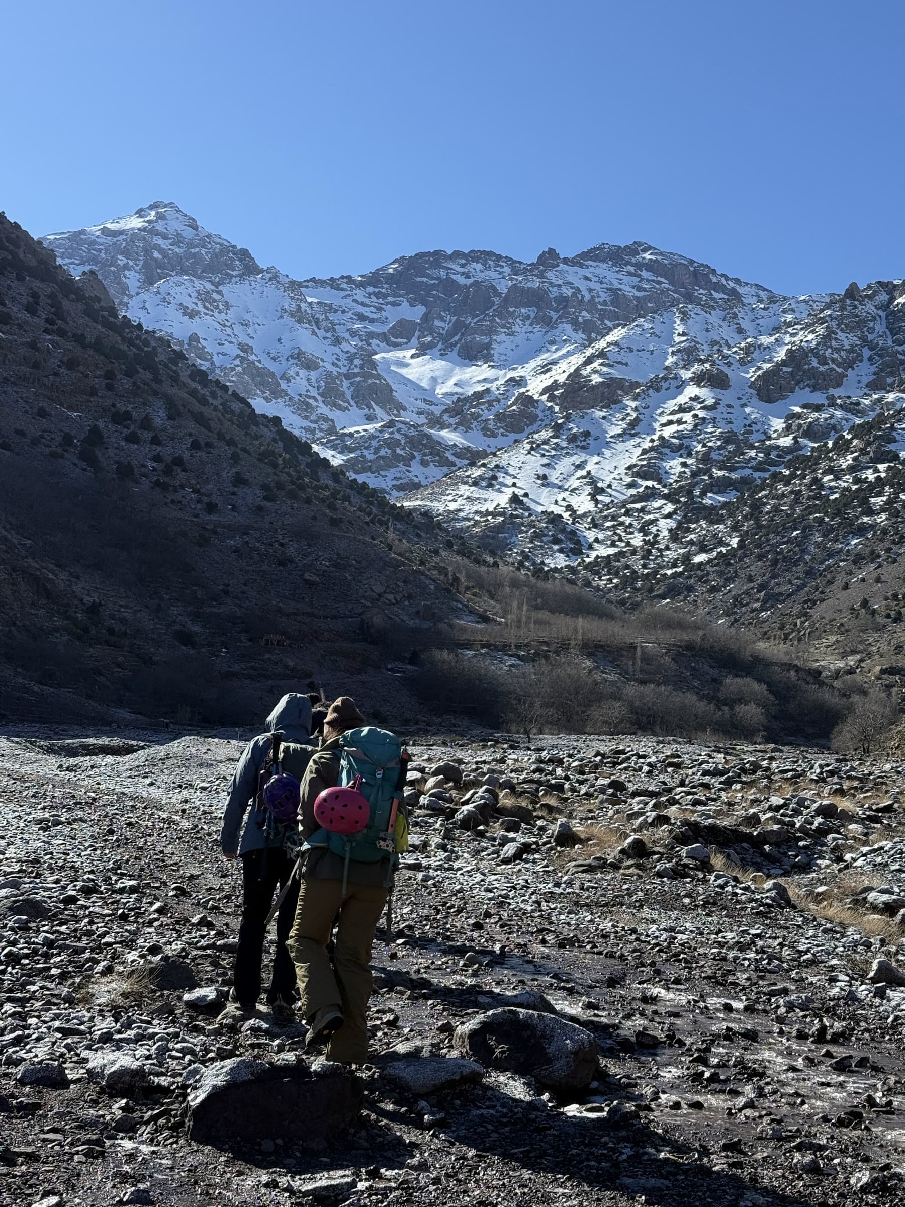 Two walkers crossing a dry boulder riverbed, the snow line and high peaks directly ahead