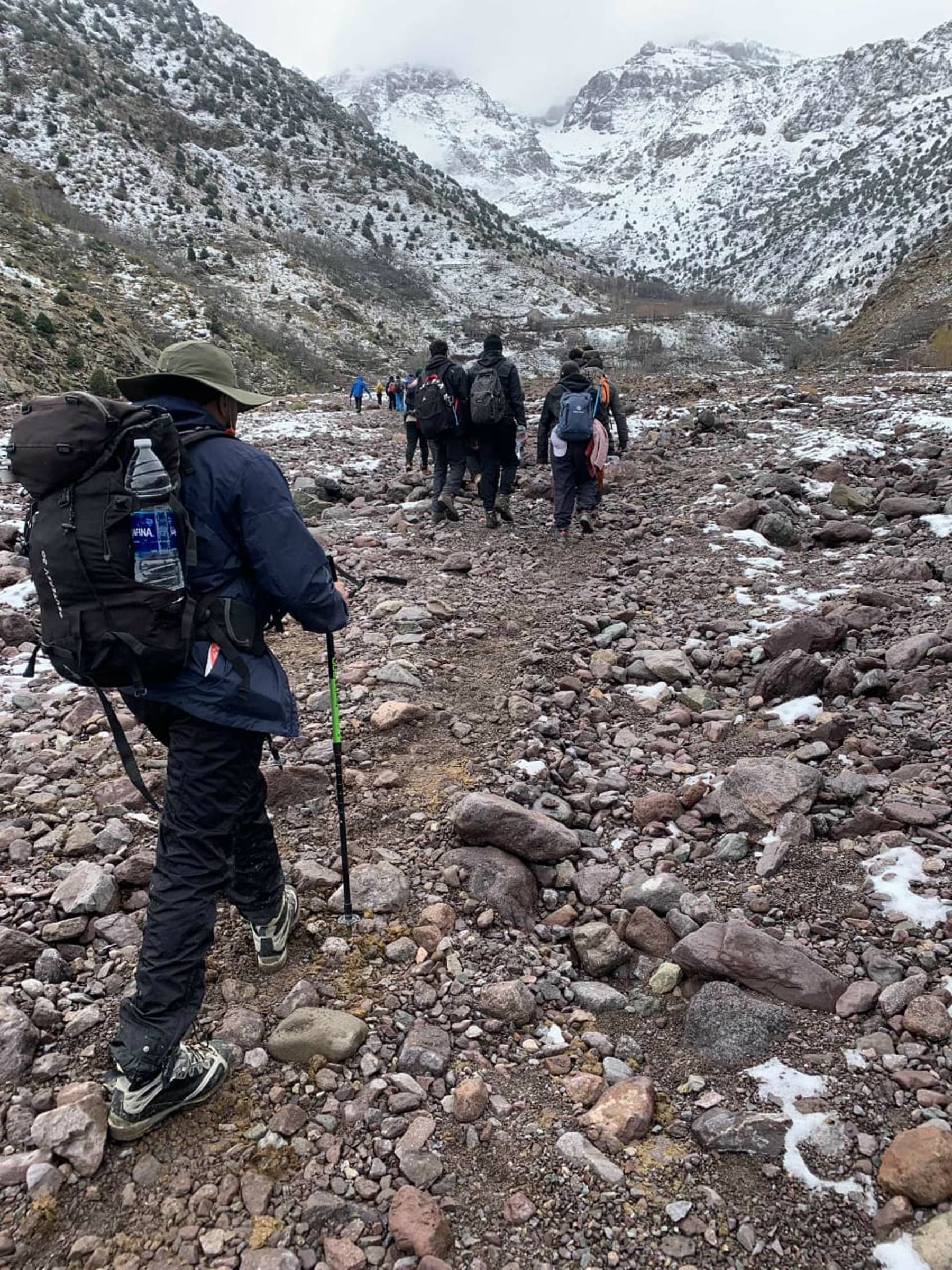 A line of walkers on the stony trail under cloud, snowed-in peaks closing the head of the valley